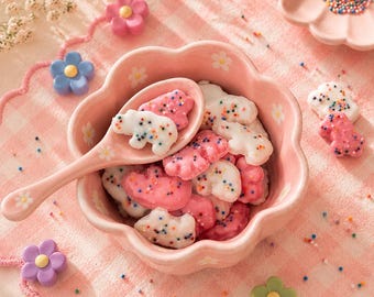 Puede incluir: Un cuenco y una cuchara rosas con forma de flor llenos de galletas con forma de oso, unas rosas y otras blancas, con chispas de colores. El cuenco está sobre un mantel individual rosa de cuadros, rodeado de decoraciones con forma de flor y un plato de chispas.