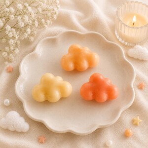 May include: Three cloud-shaped soaps in shades of yellow, orange, and coral are arranged on a scalloped plate. A lit candle in a glass holder and decorative stars and clouds are also in the image, creating a soft, pastel aesthetic.