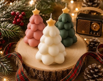May include: Three Christmas tree-shaped candles in pink, white, and green, each topped with a yellow star. The candles sit on a wooden slice, surrounded by pine needles, berries, and a plaid ribbon. A vintage radio and pine cones are in the background.