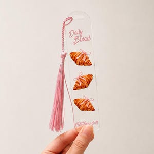 May include: Clear acrylic bookmark with a pink tassel. The bookmark features three watercolor croissants with pink bows and the text "Daily Bread" and "Matthew 6:11".