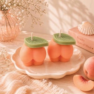 May include: Two peach-shaped candles with green leaf tops sit on a scalloped plate. The candles are orange with a peach-like texture. A book with the text "Peachy State of Mind" and a seashell are in the background.