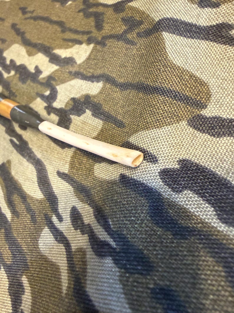 Puede incluir: Una pipa de fumar de madera con una boquilla de color claro y un v&aacute;stago de color oscuro. La pipa descansa sobre un fondo de tela de camuflaje. La pipa es de dise&ntilde;o tradicional, con forma cil&iacute;ndrica.