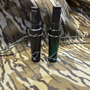 May include: Two hunting calls, one black and one dark green, with a bird silhouette design. Each call has a black mouthpiece and is attached to a camouflage lanyard. The calls are cylindrical and rest on a camouflage patterned surface.