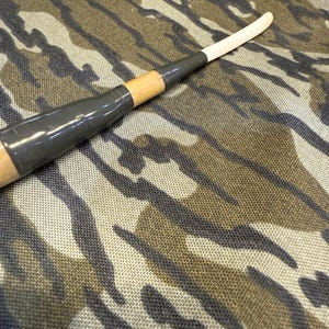 Puede incluir: Un palo de hockey de madera con una hoja blanca y un agarre negro. El palo est&aacute; envuelto con cinta gris y se coloca sobre un fondo de camuflaje.