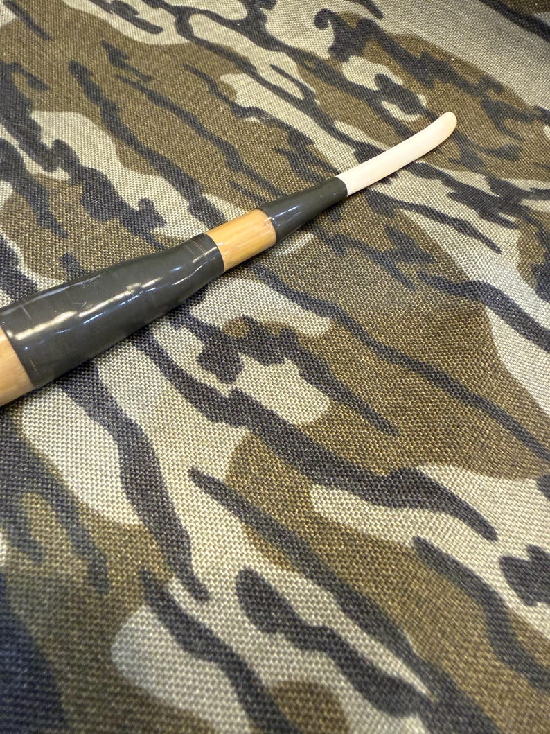 Puede incluir: Un palo de hockey de madera con una hoja blanca y un agarre negro. El palo est&aacute; envuelto con cinta gris y se coloca sobre un fondo de camuflaje.