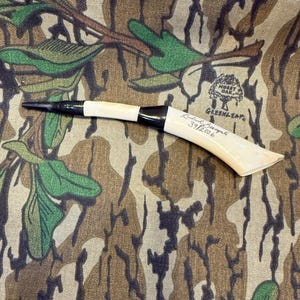 May include: A hunting call with a black tip, cream-coloured body, and a black and white accent. The call is signed and numbered, set against a camouflage fabric background.