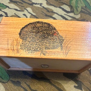May include: A rectangular wooden box featuring a dark, detailed wood-burned illustration of a turkey on the top lid. The box has a light-colored wooden frame and a small, centered circular metal pull handle on the front panel of the container.