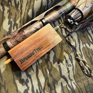 May include: A wooden hunting call with the text "Breaze Trail" engraved on it. The call is light brown and rectangular, with a small wooden peg extending from one end. A camouflage-patterned object is in the background.