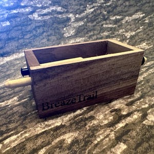 May include: A rectangular wooden box with the text "Breaze Trail" printed on the side. The box has a light brown wooden dowel and a black knob attached to the side. The box is made of dark brown wood.