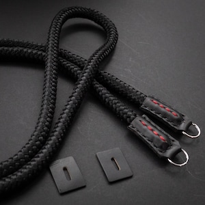 May include: Black camera strap made of braided rope with leather ends and red stitching. The strap has two metal rings for attaching to a camera.