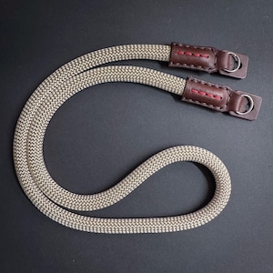 Puede incluir: Una correa de cámara beige con extremos de cuero marrón y anillos metálicos plateados. La correa está hecha de un material de cuerda trenzada.