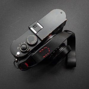 Peut inclure: Un appareil photo noir avec une sangle en cuir et un accent rouge. L'appareil photo a un cadran avec des chiffres et un petit bouton rouge. L'appareil photo est sur une surface noire.