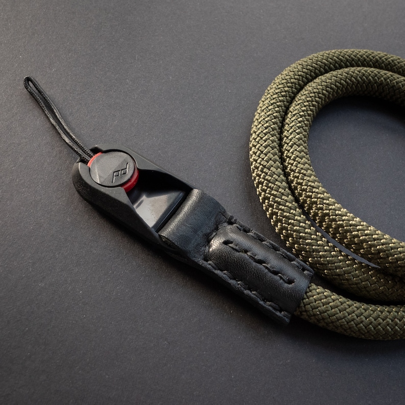 Op de afbeelding: Een zwarte en groene cameraband met een zwarte leren band en een zwarte plastic clip met een rode knop. De band is gemaakt van geweven materiaal.
