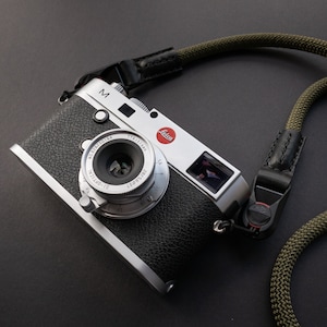 Op de afbeelding: Een zilveren Leica M camera met een zwarte leren riem. De camera heeft een zilveren lens met een zwarte lensdop. De camera heeft een rood "Leica" logo bovenop.