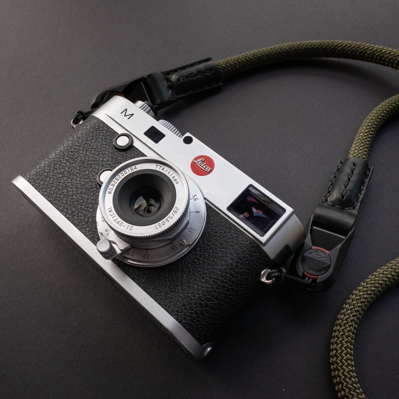 Op de afbeelding: Een zilveren Leica M camera met een zwarte leren riem. De camera heeft een zilveren lens met een zwarte lensdop. De camera heeft een rood "Leica" logo bovenop.