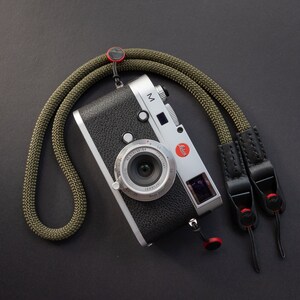 Op de afbeelding: Een zilveren camera met een zwarte leren band en een groene cameraband. De camera heeft een grote lens en een klein LCD-scherm. De camera ligt op een zwart oppervlak.