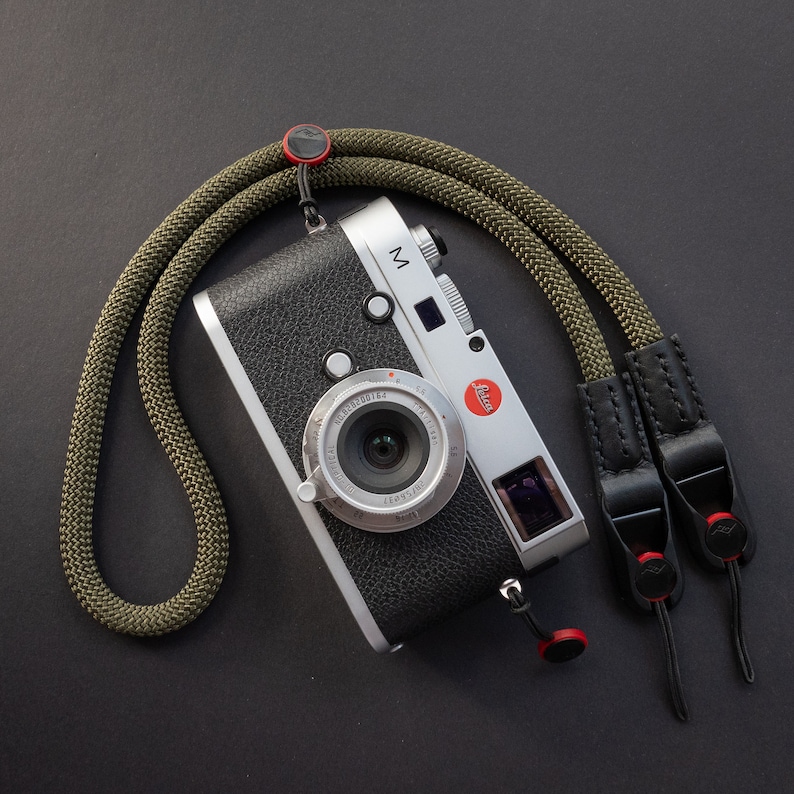 Op de afbeelding: Een zilveren camera met een zwarte leren band en een groene cameraband. De camera heeft een grote lens en een klein LCD-scherm. De camera ligt op een zwart oppervlak.