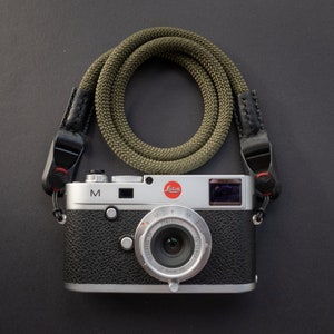 Op de afbeelding: Een zilveren Leica M camera met een zwarte leren band en een groene gevlochten cameraband. De camera heeft een zwart leren body en een zilveren lens.