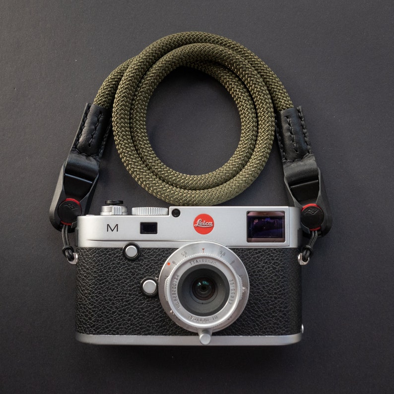 Op de afbeelding: Een zilveren Leica M camera met een zwarte leren band en een groene gevlochten cameraband. De camera heeft een zwart leren body en een zilveren lens.