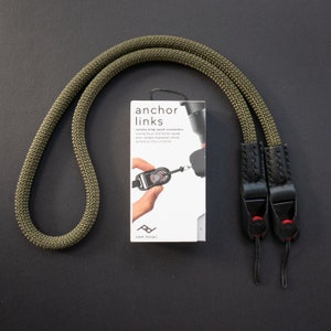 Op de afbeelding: Een groene cameraband met zwarte leren uiteinden en snelontkoppelingsconnectoren. De band is gemaakt door Peak Design en is gelabeld "anchor links".