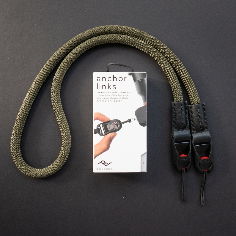 Op de afbeelding: Een groene cameraband met zwarte leren uiteinden en snelontkoppelingsconnectoren. De band is gemaakt door Peak Design en is gelabeld "anchor links".