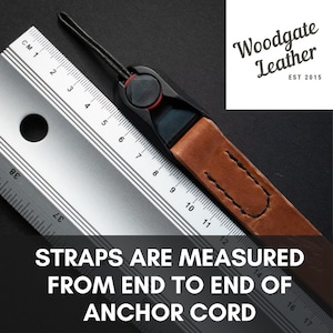 Op de afbeelding: Een bruine leren cameraband met een zwarte verstelbare gesp en een zwart koord. De band wordt van eind tot eind van het ankerkoord gemeten. De band ligt op een zilveren liniaal met de tekst "Woodgate Leather EST 2015" op de achtergrond.