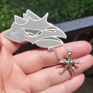 Può includere: Uno spillo in smalto argentato con la testa di un uccello grigio stilizzata con una maschera nera e una catena argentata attaccata a un ciondolo a forma di stella argentato.