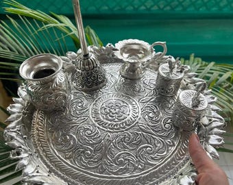 Peut inclure: Un ensemble de puja thali argenté avec des motifs floraux et de feuilles complexes. L'ensemble comprend un grand plateau rond, de petites tasses, un récipient et un support orné. Le bord du plateau est décoré d'un motif de pétales.