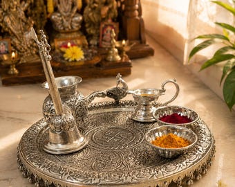 May include: A silver-toned metal puja thali set featuring an embossed circular tray with a central swastika symbol, a bell, a peacock-shaped oil lamp, and two small bowls containing red and yellow powders, all with intricate floral detailing.