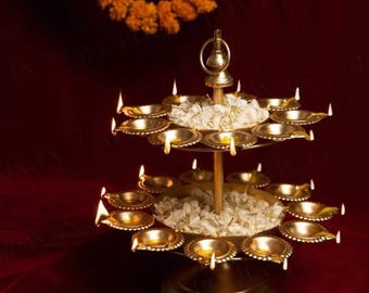 May include: A two-tiered brass diya stand with multiple small bowls for oil lamps. The stand is decorated with white flower petals and lit with small flames.