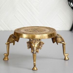 Peut inclure: Un support décoratif en métal doré avec un dessus rond et trois pieds en forme d'éléphants. Le support présente des détails complexes et un motif sur le dessus.