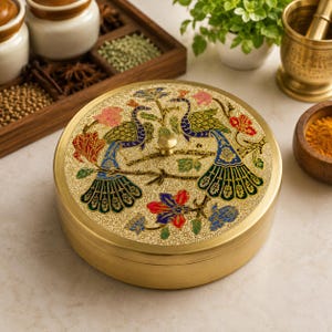 Op de afbeelding: Een ronde opbergdoos van messing met een plat deksel voorzien van een gedetailleerde, kleurrijke emaille inleg van twee pauwen op takken omringd door bloemmotieven. De doos heeft een gladde, geborstelde gouden afwerking en een kleine knop.