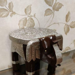 May include: A wooden elephant stool with a silver-coloured top and legs. The stool has a carved elephant head and ears.