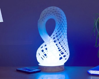 Könnte beinhalten: Eine moderne weiße Lampe mit einem einzigartigen, offenen Design, das ein sanftes Leuchten ausstrahlt. Die Lampe steht auf einem Holzsockel und wirft eine blaue Reflexion auf die darunter liegende Oberfläche. Ein Smartphone und ein blaues Buch liegen ebenfalls auf dem Tisch.