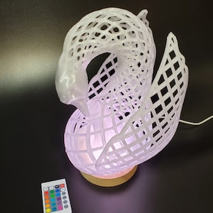 Può includere: Una lampada bianca a forma di cigno con un design a griglia. La lampada poggia su una base di legno ed emette un bagliore morbido e colorato. Un telecomando con opzioni di colore è accanto alla lampada. La lampada è collegata con un cavo bianco.