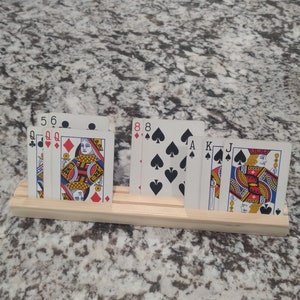 May include: A wooden card holder with six playing cards. The cards are a Queen of Hearts, 5 of Clubs, 6 of Clubs, 8 of Spades, Ace of Spades, Jack of Spades, and King of Spades.