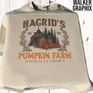 May include: A beige sweatshirt with a graphic design of a rustic cabin with a pumpkin patch. The text reads "Hagrid's Pumpkin Farm Magically Grown" with the dates "Est. 1981".