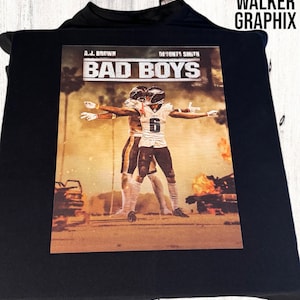 以下が含まれることがあります： 炎の爆発を背景にした、2人のアメリカンフットボール選手がアクションを起こしているグラフィックプリントが施された黒いTシャツ。グラフィックには「Bad Boys」というテキストと、その上に「A.J. Brown」と「DeVonta Smith」の名前が書かれている。