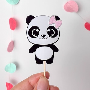 May include: A black and white panda bear cupcake topper with a pink bow. The topper is on a wooden stick and is ready to be placed on a cupcake.