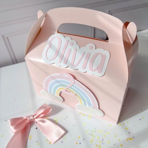 May include: Pink favor box with a rainbow and clouds design. The box is personalized with the name "Olivia" and the number "2".