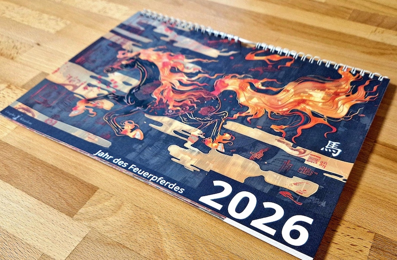 May include: A spiral-bound calendar for 2026 featuring a stylized illustration of a fiery horse in shades of orange, red, and black against a dark blue background. The calendar has text in German and Japanese.