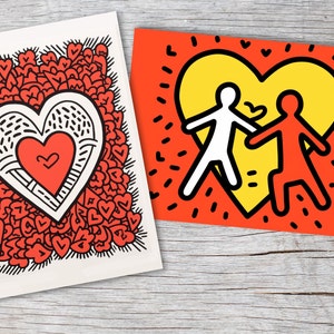 Puede incluir: Dos impresiones art&iacute;sticas con dise&ntilde;os de coraz&oacute;n. Una presenta un gran coraz&oacute;n lleno de corazones m&aacute;s peque&ntilde;os, y la otra muestra dos figuras tomadas de la mano dentro de un coraz&oacute;n amarillo sobre un fondo rojo. Ambas est&aacute;n sobre una superficie de madera.