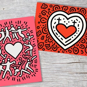 Puede incluir: Dos impresiones art&iacute;sticas con dise&ntilde;os gr&aacute;ficos llamativos. Una es rosa con figuras blancas que rodean un coraz&oacute;n blanco. La otra es roja con un dise&ntilde;o de coraz&oacute;n, contornos negros y elementos decorativos. Ambas est&aacute;n sobre una superficie de madera.