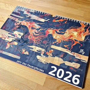 May include: A spiral-bound calendar for 2026 featuring a stylized illustration of a running horse with fiery orange and red mane and tail. The calendar has a dark blue background with Japanese text and the words "Jahr des Feuerpferdes".
