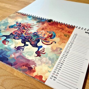 May include: A spiral-bound calendar open to March 2026, featuring a colorful illustration of a horse with a flowing mane and tail. The horse is depicted in shades of blue, purple, orange, and red against a cloudy background. The word "M&auml;rz" is visible.