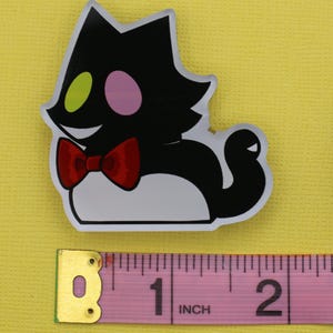 Puede incluir: Una pegatina con forma de gato negro con vientre blanco, pajarita roja y cara sonriente. El gato tiene un ojo verde y un ojo rosa. La pegatina mide aproximadamente 5 cm de largo.