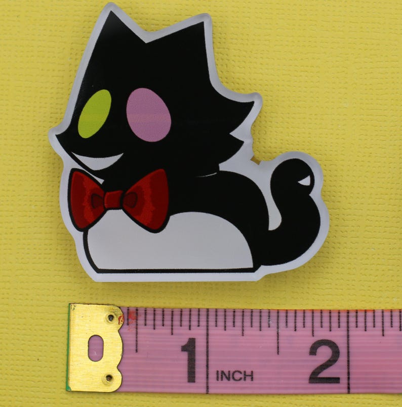 Puede incluir: Una pegatina con forma de gato negro con vientre blanco, pajarita roja y cara sonriente. El gato tiene un ojo verde y un ojo rosa. La pegatina mide aproximadamente 5 cm de largo.