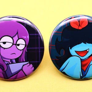 K&ouml;nnte beinhalten: Zwei runde Ansteckbuttons mit Cartoon-Illustrationen. Der linke Button zeigt eine violetth&auml;utige Figur mit Brille, die einen grauen Gegenstand h&auml;lt. Der rechte Button zeigt eine blauh&auml;utige Figur mit schwarzen Haaren, roter Schleife und rotem Schal.