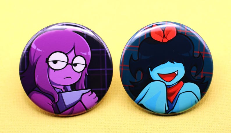 K&ouml;nnte beinhalten: Zwei runde Ansteckbuttons mit Cartoon-Illustrationen. Der linke Button zeigt eine violetth&auml;utige Figur mit Brille, die einen grauen Gegenstand h&auml;lt. Der rechte Button zeigt eine blauh&auml;utige Figur mit schwarzen Haaren, roter Schleife und rotem Schal.