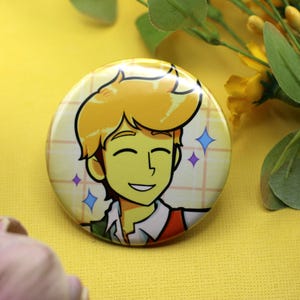 K&ouml;nnte beinhalten: Ein runder Ansteckbutton mit einer Cartoon-Illustration einer l&auml;chelnden Person mit blonden Haaren, die ein wei&szlig;es Hemd mit Kragen und eine rote Weste tr&auml;gt. Der Hintergrund der Illustration ist ein hellgelbes Gittermuster mit kleinen Sternsymbolen.